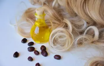 A jar of jojoba oil and spread out seeds beside locks of healthy hair