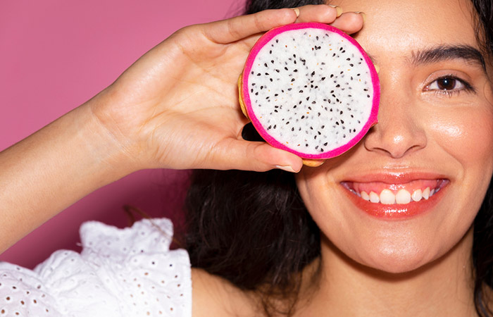 Woman with radiant skin smiles as she covers one eye with a slice of dragon fruit