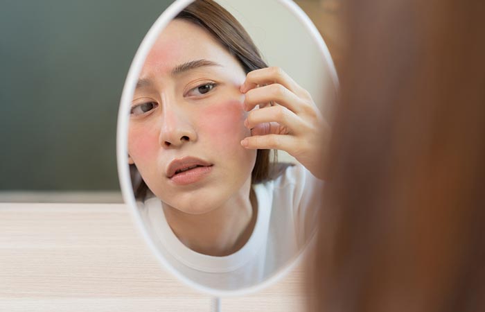 A young woman looks into the mirror with a red patch or rash on her face.
