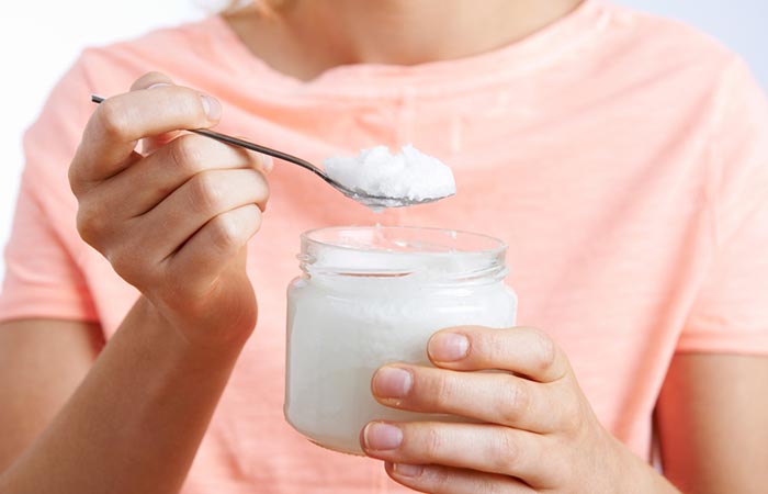 A woman holds a spoonful of coconut oil scooped out of a jar as a remedy for dry scalp