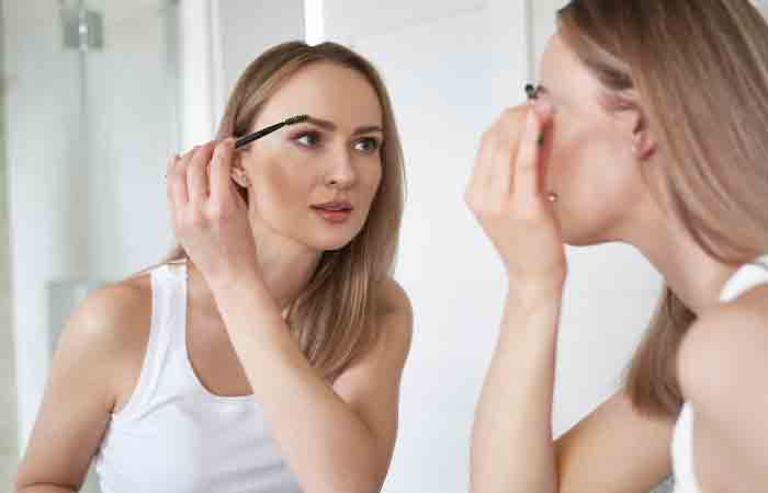 Woman looking in the mirror and applying mascara on her gray eyerbrows.