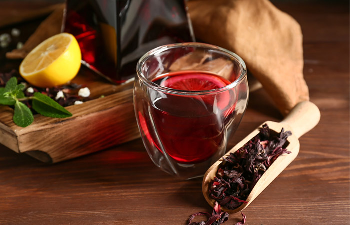 A cup of fresh hibiscus tea with a scoop of dried hibiscus petals and other ingredients in the background