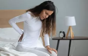 Woman in discomfort while holding her back and sitting on the bed