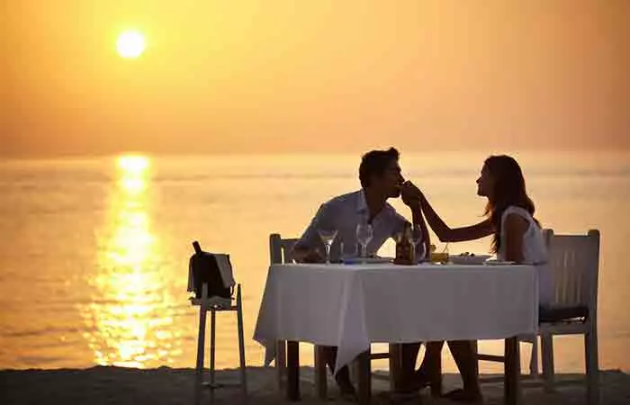 Man kissing woman's hand at dinner table against a beach sunset background