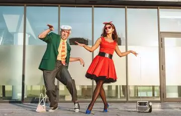 older man and younger woman dressed in costumes do a quirky dance on a city pavement