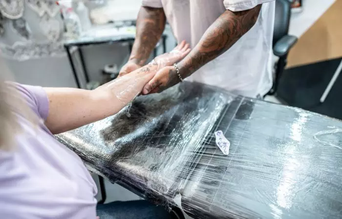 A tattoo artist covers a woman’s tattoo with Saniderm plastic wrap at a tattoo studio