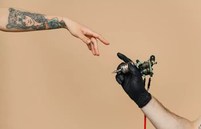 A tattooed hand and a gloved hand with tattoo equipment reaching towards each other