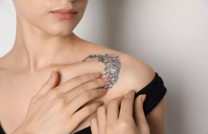 Bust of a woman applying Lubriderm cream on a tattoo on her shoulder.