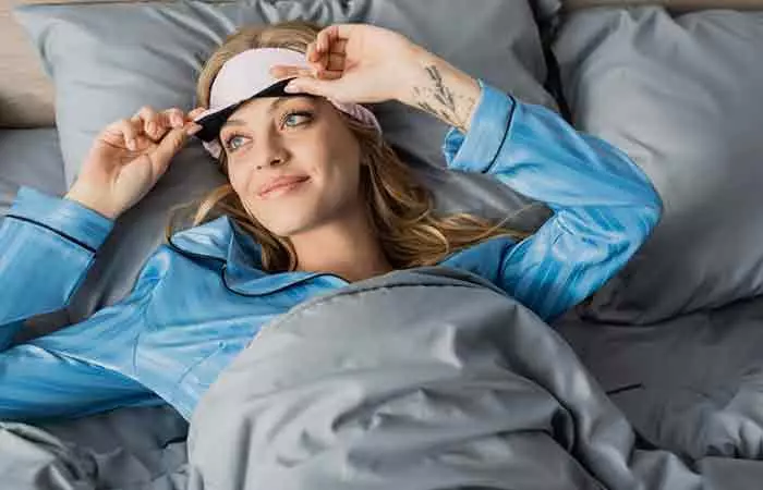 A woman with tattoos on her hand adjusts her sleeping mask in bed.