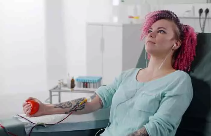A tattooed woman lying back and pumping a rubber ball while donating blood at a medical center