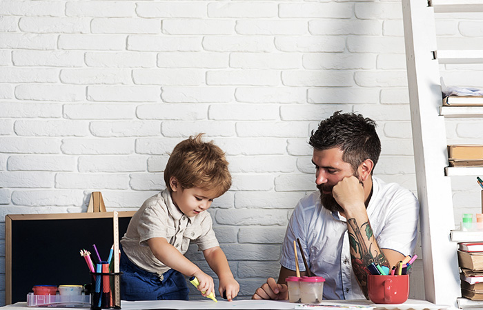 A tattooed teacher looks on as his young student draws on a piece of paper