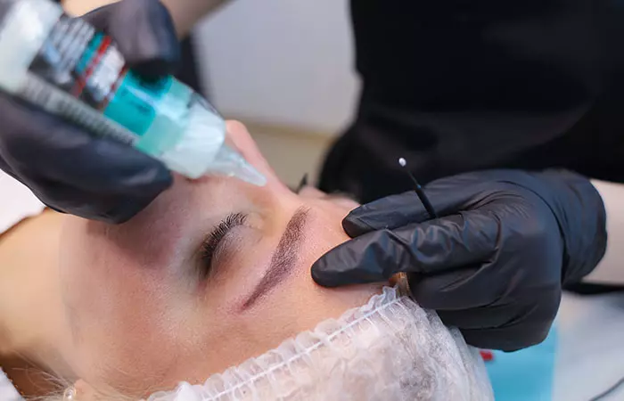 An aesthetician applying a topical anesthetic to the patient’s eyebrow before microblading