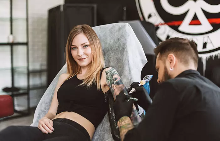 A woman looks on as a tattoo artist performs a tattoo cover-up on her upper arm