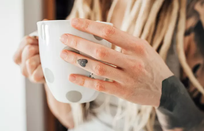 Woman with a faded heart tattoo on her middle finger, holding a cup