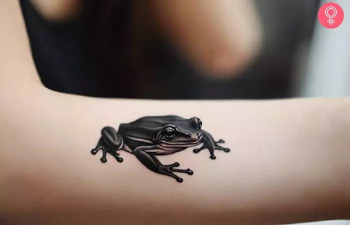Black frog tattoo on a woman’s forearm